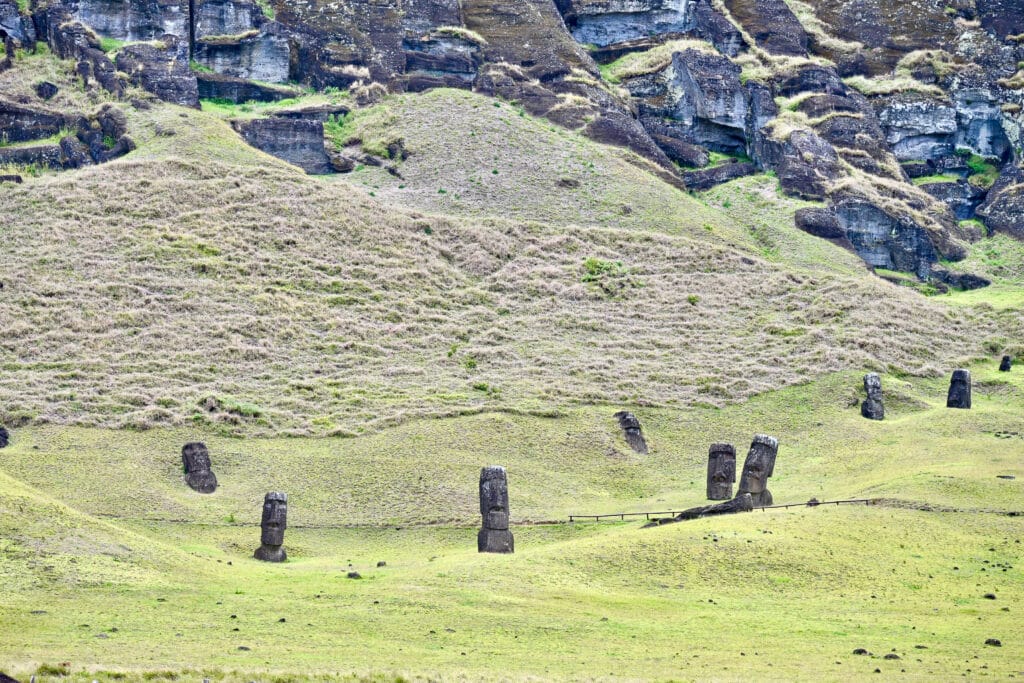 Pale green hilly grass patch leading up to a rocky hillside. It is scattered with large stone human form statues at the bottom. They are leaning in different directions.