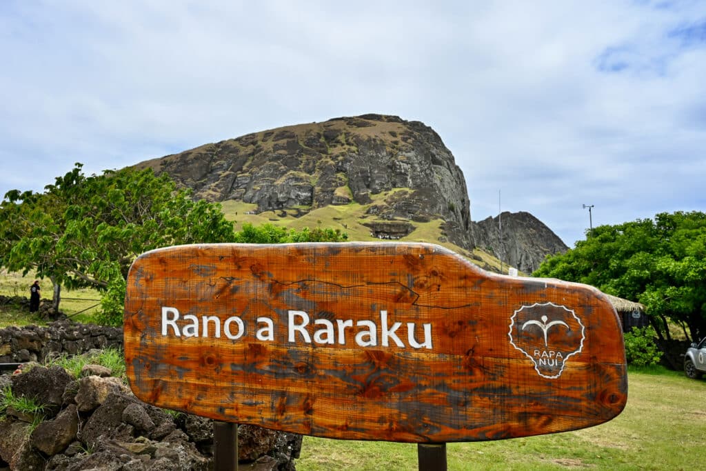 A wooden sign post with the words Rano Raraku stands in the front of the frame. A rocky peak behind. Grass in the foreground. Pale blue cloudy sky above