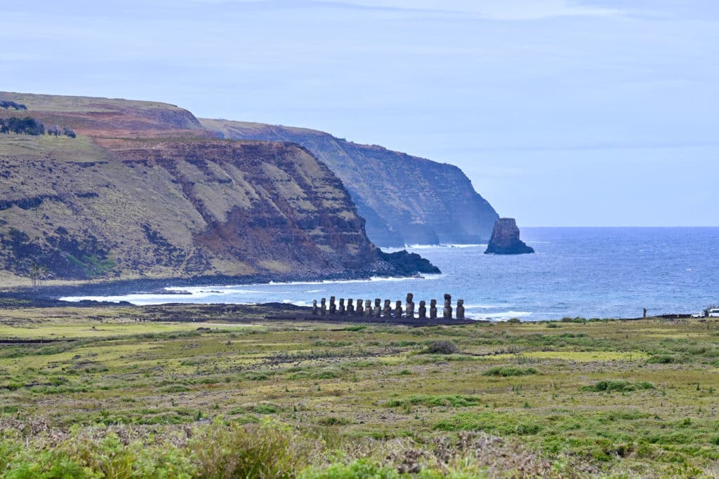 Looking down a grassy hill to a coastal bay surrounded by cliffs. On the edge of bay is a wide platform with standing statues overlooking the water
