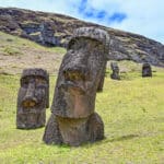 A grassy slope bottom left with a white cloudy sky above. Two large stone figure heads stand on the grass. The larger one to the right hand bottom corner is leaning to the right. The smaller one to the left hand side is more upright. Deep seat eye sockets and long nose