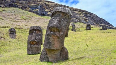 A grassy slope bottom left with a white cloudy sky above. Two large stone figure heads stand on the grass. The larger one to the right hand bottom corner is leaning to the right. The smaller one to the left hand side is more upright. Deep seat eye sockets and long nose