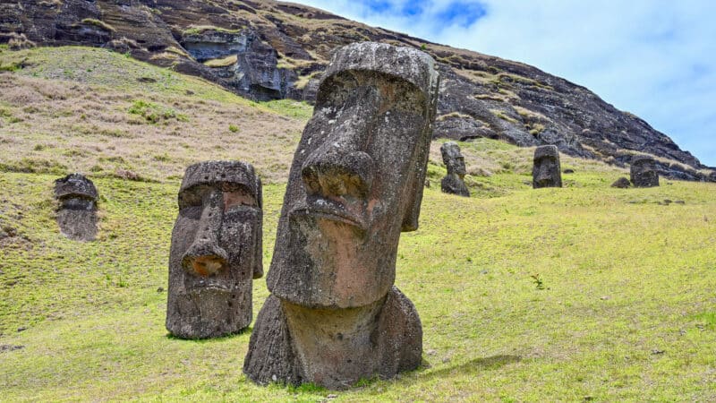 A grassy slope bottom left with a white cloudy sky above. Two large stone figure heads stand on the grass. The larger one to the right hand bottom corner is leaning to the right. The smaller one to the left hand side is more upright. Deep seat eye sockets and long nose