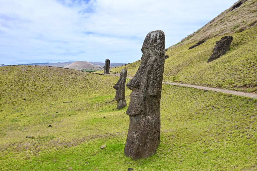 A grassy bank with cloudy sky above. In the foreground is a tall stone carving of a human figure (head and torso) sideways to the camera. It has a long face. There is a second one behind it to the left and third on on the hill in the background