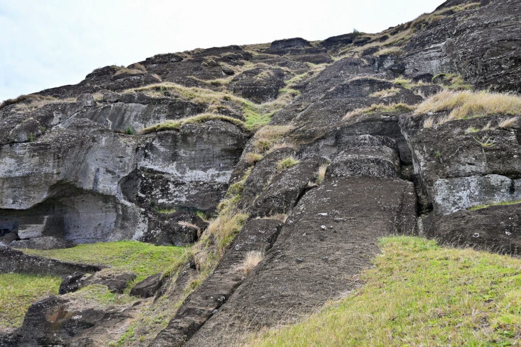 Scene of a stone quarry wall with grey sky above. Evidence of a figure statue lying on the ground to the right hand side