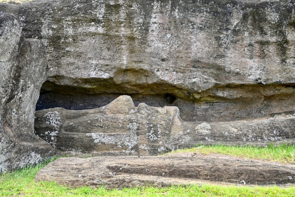 View of a stone wall at the top, with a large stone figure carving lying on its back at the bottom on grass