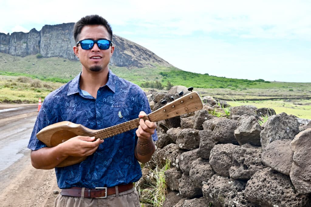 A man in a blue shirt and blue sunglasses stands to the left of the frame holding a ukulele. There is a stone wall to his right and fields behind him leading to a rocky peak. Cloudy sky above.