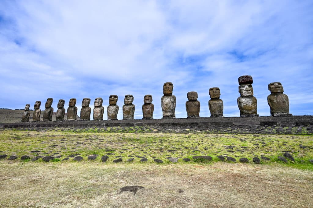 Grassy back with a wide stone platform stretching across the frame supporting 15 human form stone carved statues - different shapes and sizes. One has a red hat. Area in front of the platform remarked with ring of stones. Blue sky with patchy cloud above.