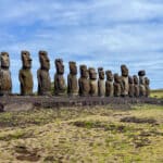 View across faded yellow grass to a wide stone platform running across the frame. Standing proudly on the platform are 15 stone statues with long heads and torsos, all different sizes. Blue sky with whispy white cloud above