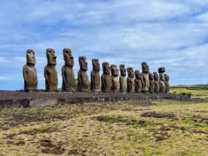 View across faded yellow grass to a wide stone platform running across the frame. Standing proudly on the platform are 15 stone statues with long heads and torsos, all different sizes. Blue sky with whispy white cloud above