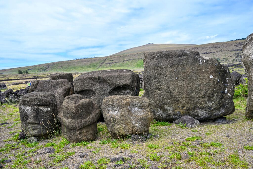 Field with patchy grass and hill behind. Pale blue sky filled with patchy cloud. Rows of carved stone blocks sit on the grass. The first three look like human faces. Larger blocks behind- eye sockets visible.