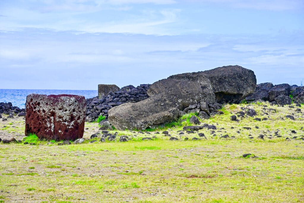 A yellowy green grass field at the bottom of the frame. Pill of stones across the horizon with a large fallen stone statue (looks like a large slab of stone). To the left there is a circular red stone covered in moss sitting on the grass. Ocean and blue sky behind.