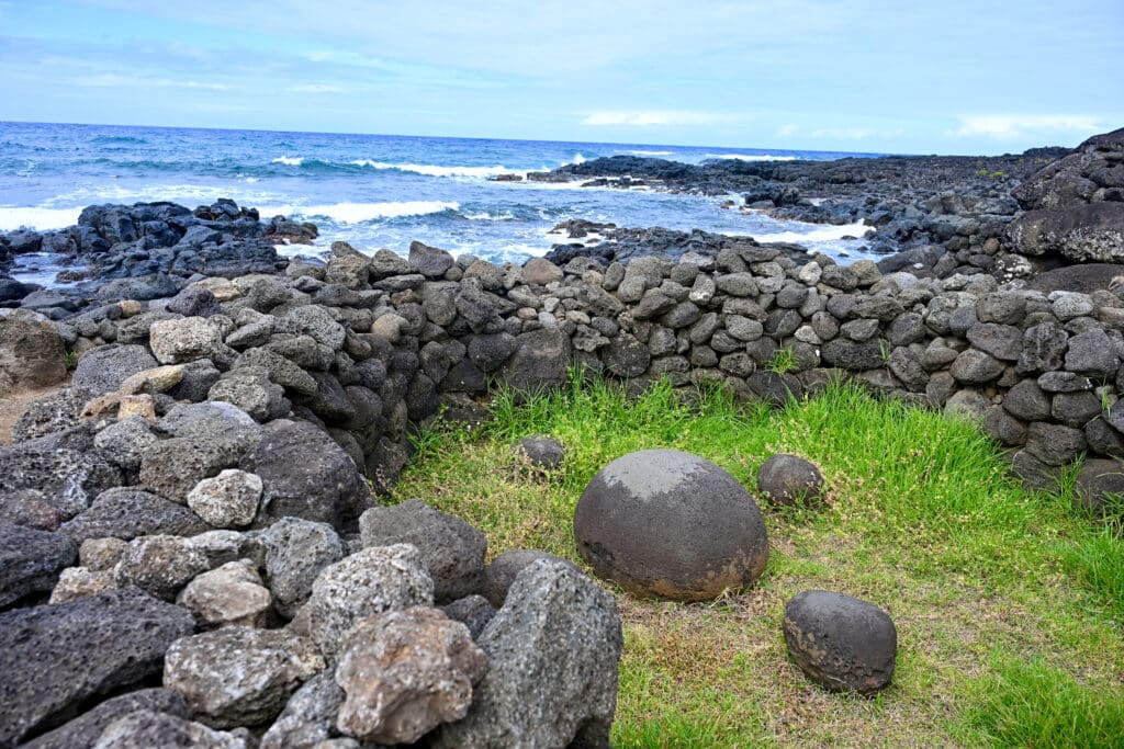 A circle of pilled dry stone walls with grass inside. In the middle is a large circular stone surrounded by 3 or 4 smaller circular stones. Rocky shore and ocean behind. Pale blue sky