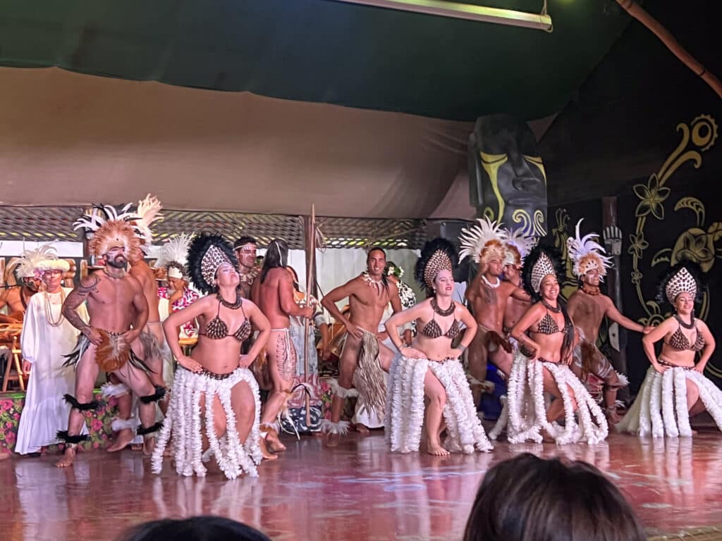 Two lines of dancers in Polynesian outfits. 4 girls in front in white flower garland skirts, bikini tops and headdresses. Male dancer in feather costumes behind.