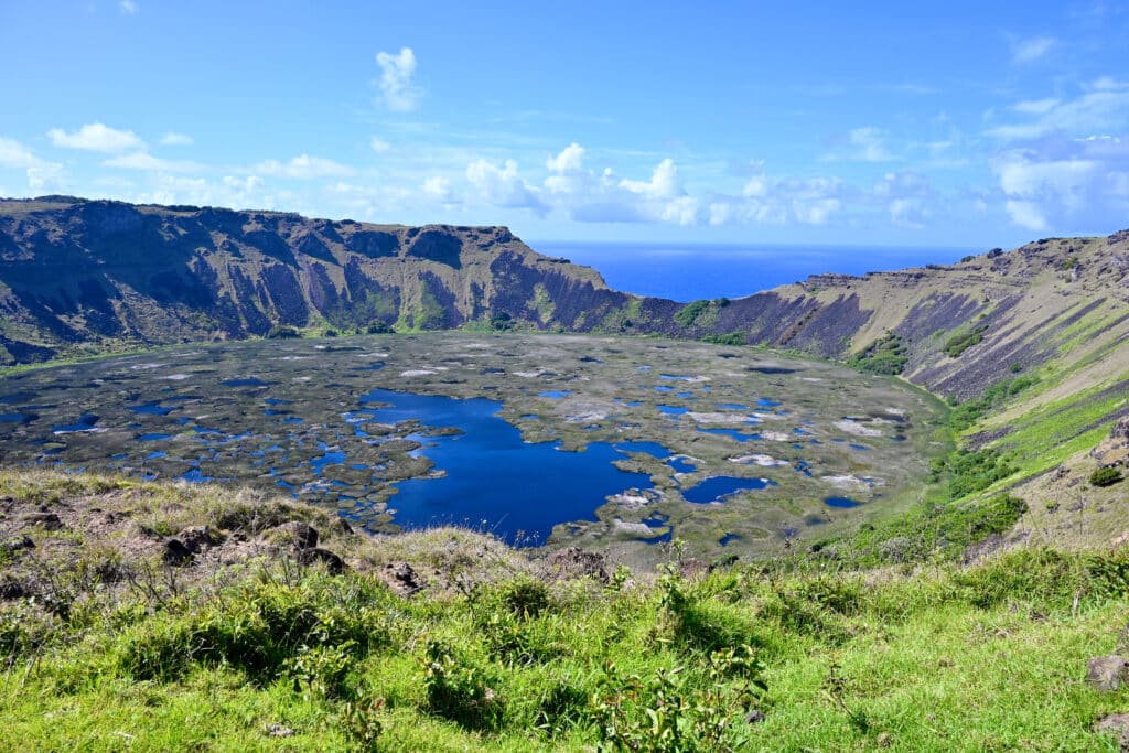 A large circular volcano crater fills the middle of the frame. It is filled with dark blue water and is covered in patches of grassy moss. Steep sides of the volcano. Grassy ledge in bottom and ocean on the horizon. Pale blue sky above.