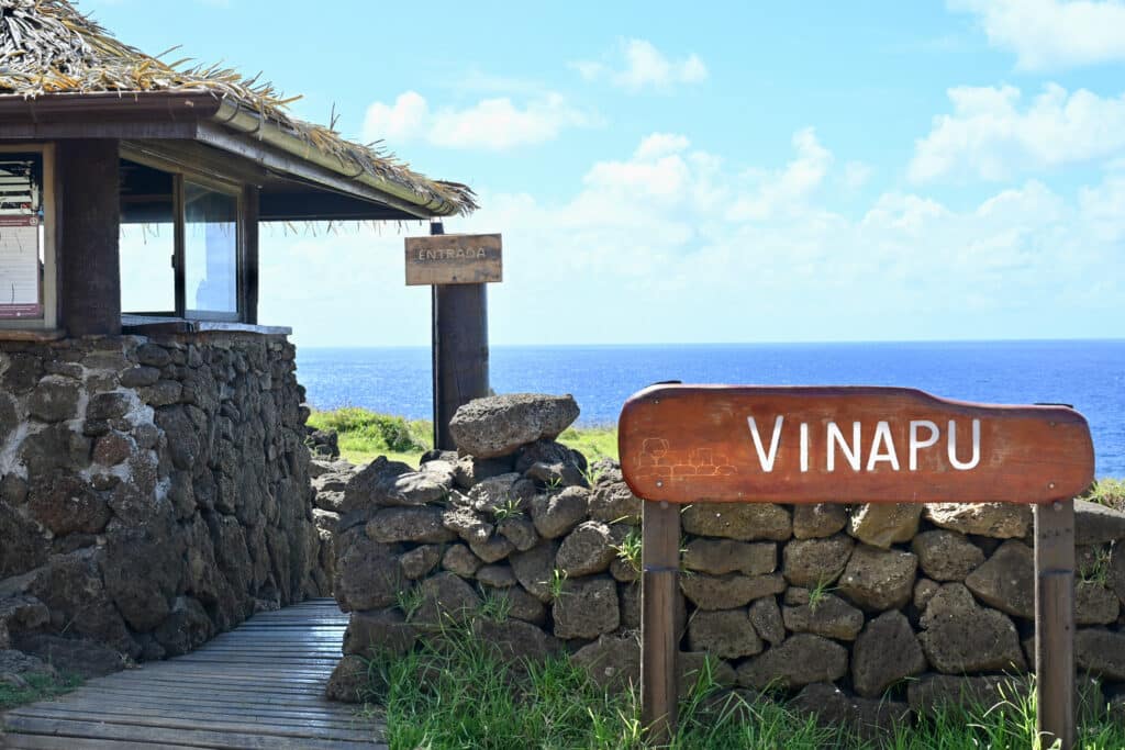 A stone wall stands to the right hand side with a wooden signpost inside saying Vinapu in white letters. A path and office to the left. Ocean and blue sky above