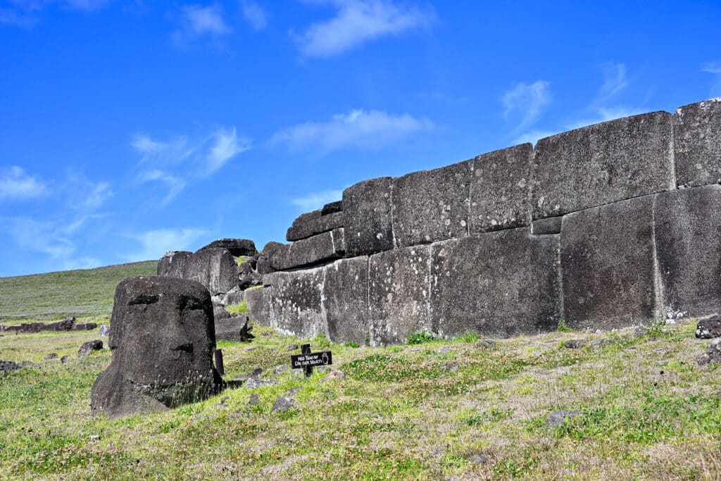looking up a pale grassy bank to a stone platform made of closely fitting blocks making a flat frontage. A stone with a carved face sits to the bottom left. Blue sky above