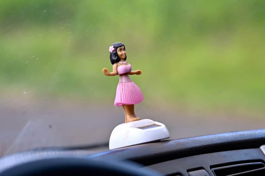 A black car dashboard and edge of steering wheel visible at bottom of frame. Green blurry background above. In the centre is a plastic Hawaiian hula girl standing on a solar panel for the car. She is wearing pink skirt, pink top and pink flower in her black hair. Air out to the left hand side