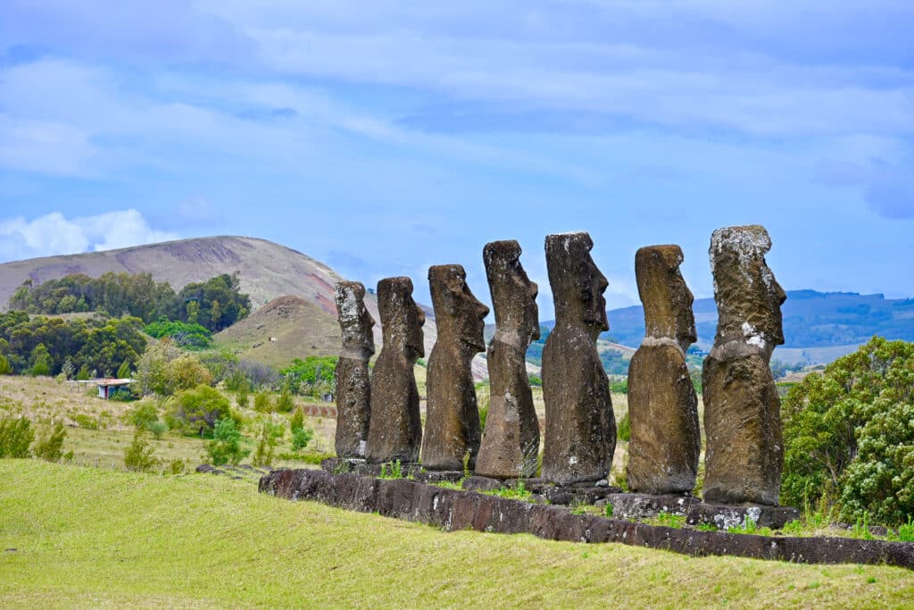 A row of 7 carved human figures stands on a platform leading from the bottom right of the frame to the middle. They are looking away from the camera. grass field to the left and hills in the background. Pale blue sky