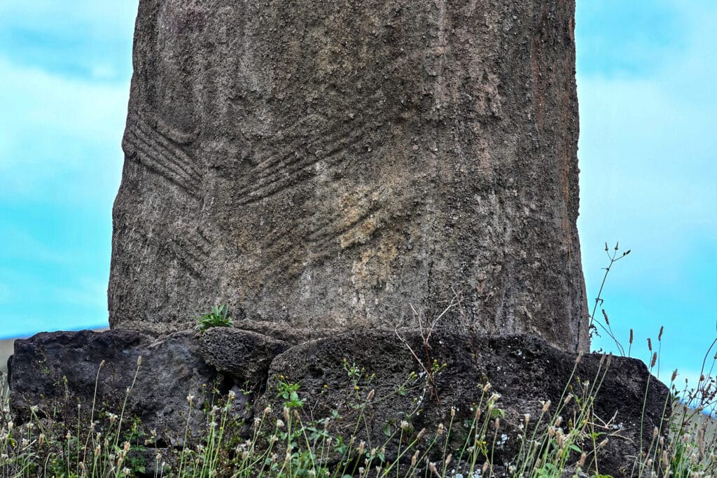 Looking up at a stone statue on a stone platform with grass plants in front. On the statue are carvings which look like fours hands - two on each side of the statue - fingers towards the middle.