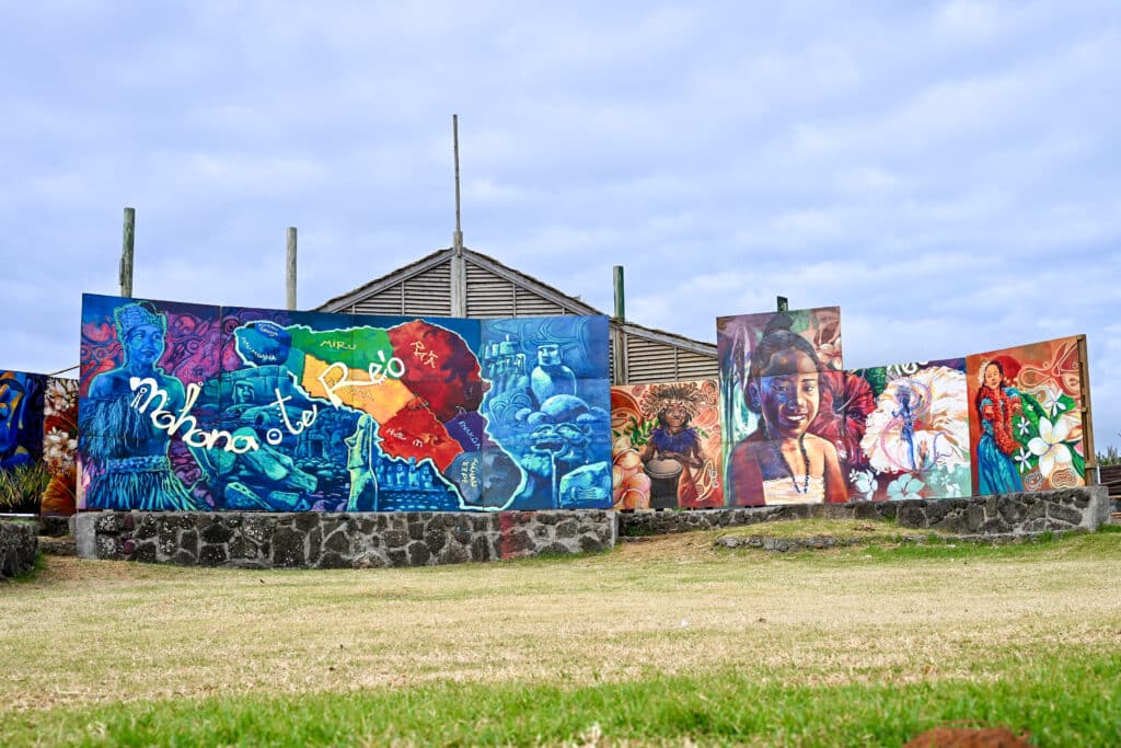 A grassy field in the bottom half of the frame. Across the horizon is a row of large painted boards featuring Polynesian designs. A building behind and cloudy sky above.
