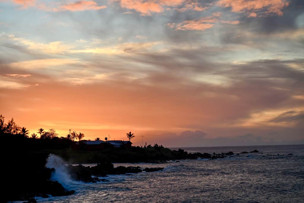 Bright orange and pink sunset over a rugged coastline silhouetted to the bottom left. Palm trees on the coast. Dark sea to the right