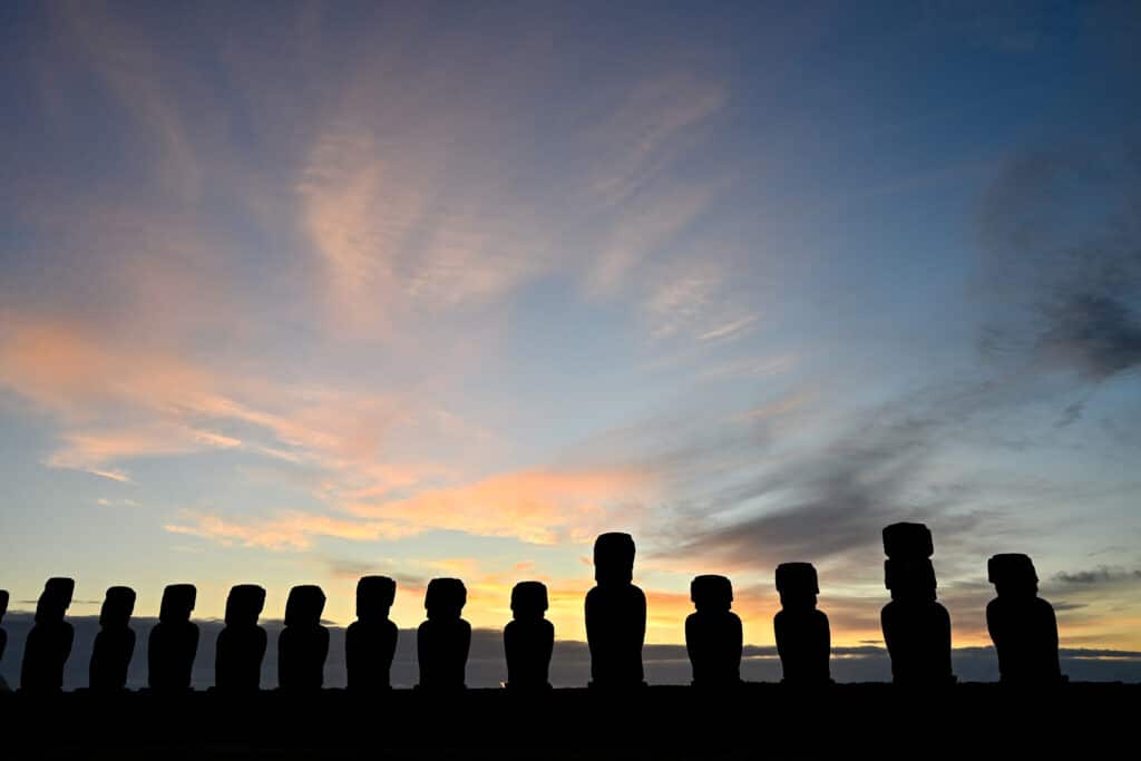 Row of 15 stone statues stretch across the bottom of the frame. they are in silhouette - all different shapes and sizes. A multi coloured sun rise sky behind.