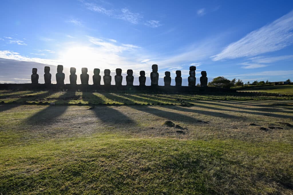 Bottom third a grassy field. A wide stone platform stretches across the middle of the frame. 15 stone carved statues stand on the platform - in human form, all different shapes and sizes. Blue sky above with whispy white cloud. Long shadows of the statues are cast on the grass in front as sunrise behind.