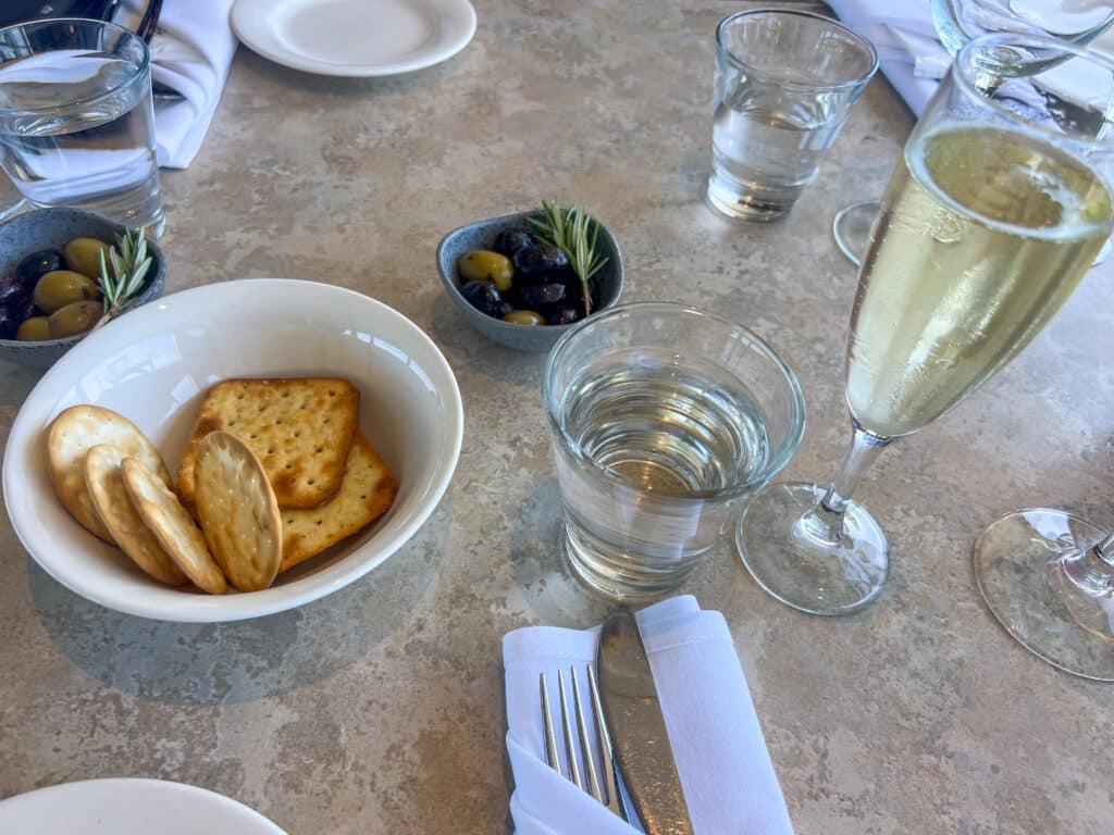 Looking down at a pale table top set for lunch. Knife and fork in a white napkin bottom centre. Small white bowl, of crackers to the left. Glass of sparkling wine to the right