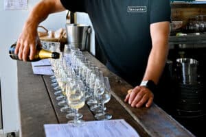 Man in black t-shirt with white logo saying Lancaster is pouring champagne from a bottom into a row of glasses lined up on a wooden bar top. His head is not shown in photo