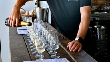Man in black t-shirt with white logo saying Lancaster is pouring champagne from a bottom into a row of glasses lined up on a wooden bar top. His head is not shown in photo