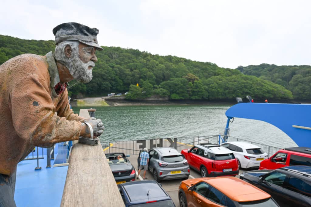 A statue of a fisherman leans on a bannister looking down on the deck of an open topped car ferry. Rows of cars lined up on the ferry as it approaches the other side of the river. Trees on the river bank. Grey cloud above.