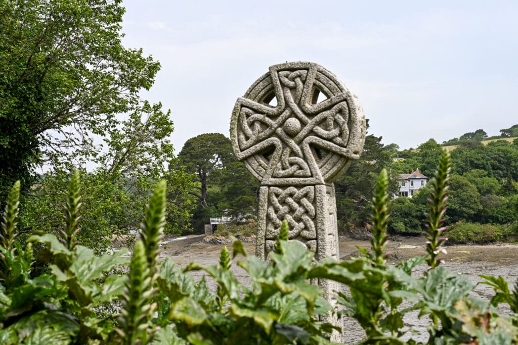 A stone Celtic cross rises up from a row of green tropical plants at the bottom. Beach behind and trees beyond that with a white houses peeping out of the trees. Pale grey sky above.
