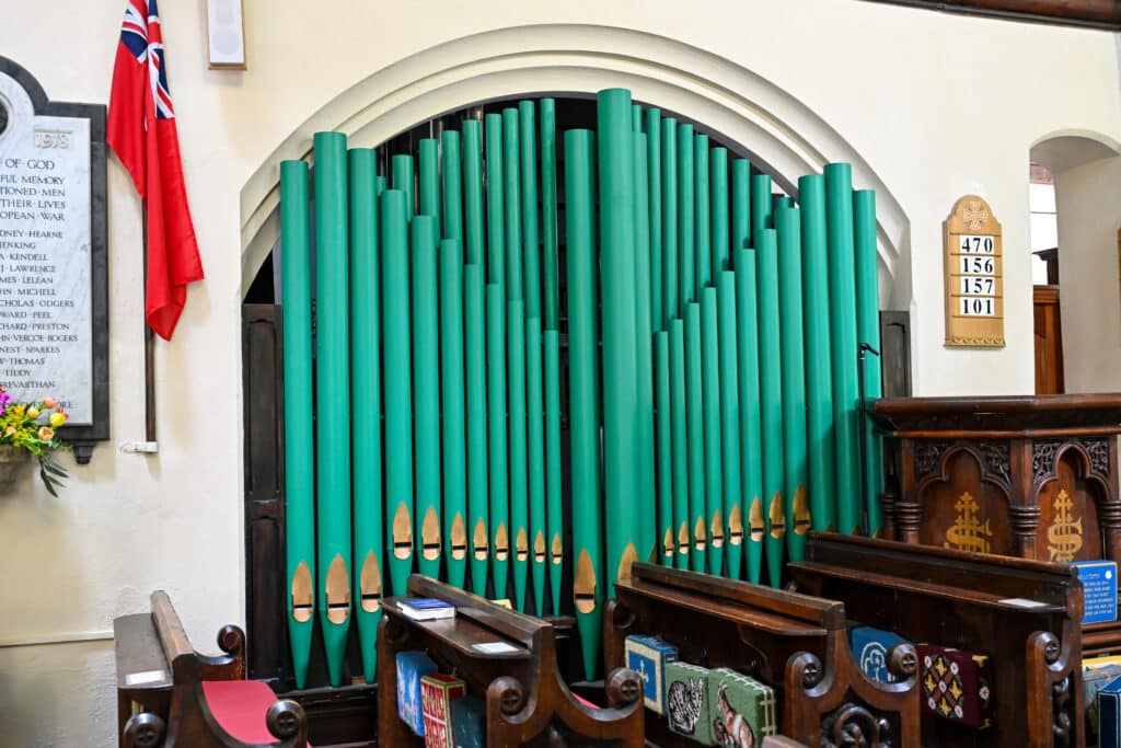 A row of turquoise organ pipes sits in an arch in the inside wall of a church wall. Wooden pews in front. Pulpit to the right.