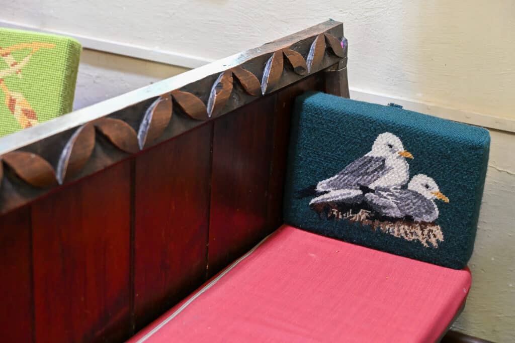 a wooden church pew sits across the middle of the frame. It has a red padded seat cushion and a jade green kneeling cushion with tapestry sea gulls on it. White wall behind.