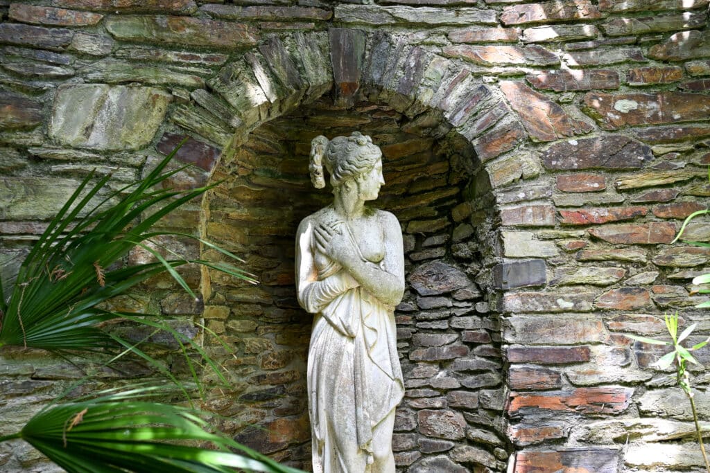 A white statue of a lady in a sheet and Roman style hair looking to the right, sits in an arched alcove of brickwork. Some green ferns to the left.
