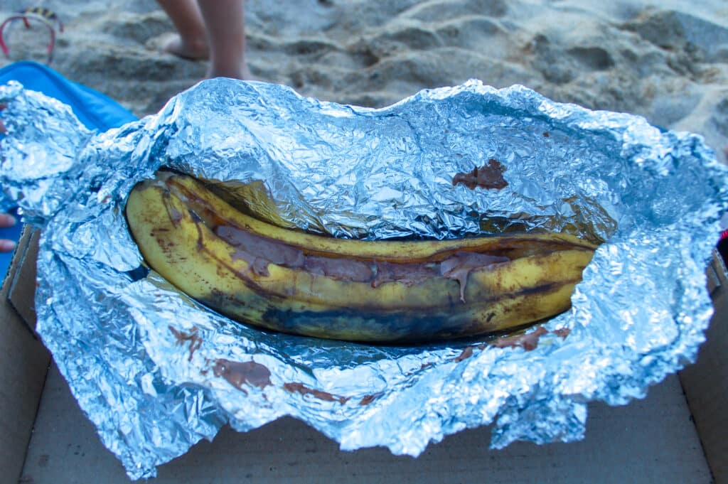 Piece of tin foil holding up a banana backed with chocolate in the middle. Beach in background