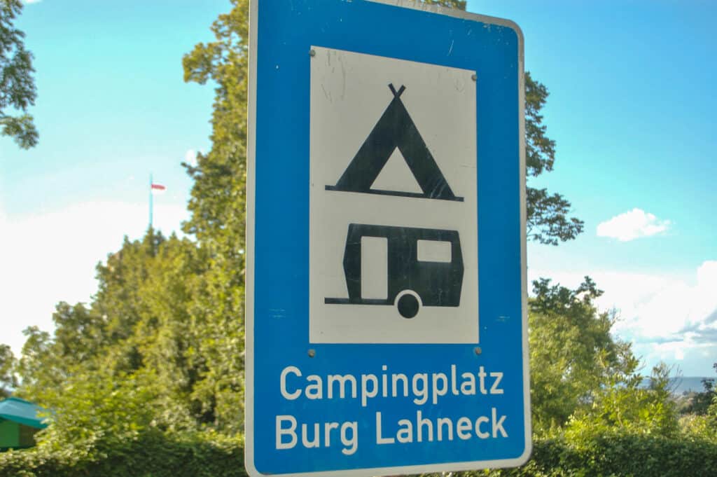 Blue sign advertising a tent and caravan site (black logos on white square). Campingplatz Burg Lahneck. Trees and pale blue sky behind