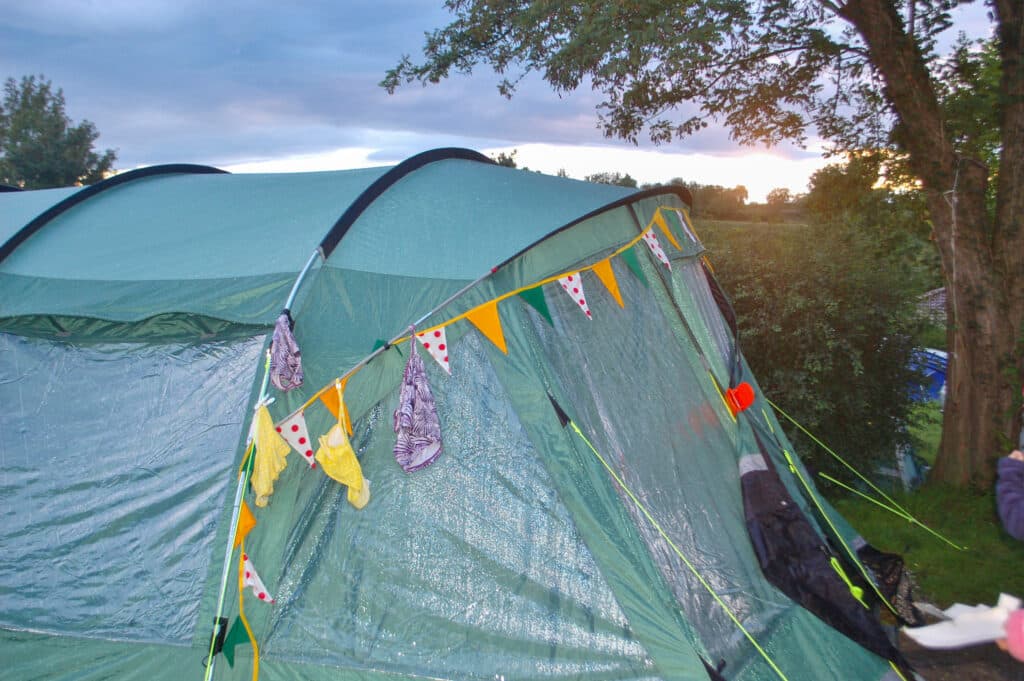 Large green tent erected to the left of the frame. It has multicoloured bunting around the front. Tree to the right. Sunset above.