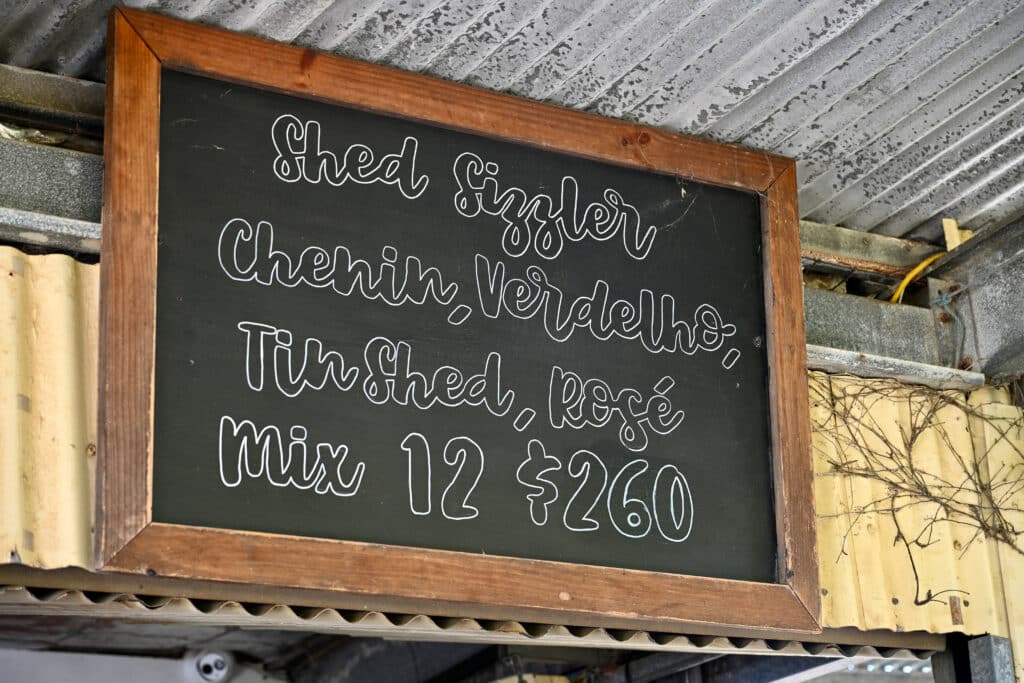 Looking at a black chalk board with wooden frame and corrugated roof. It has the name of wines chalked on the sign