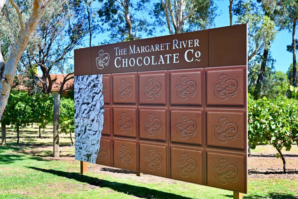 A large signpost sits across the middle of the frame on grass with blue sky and tall trees behind. It advertises the Margaret River Chocolate Company above a visual of squares of chocolate
