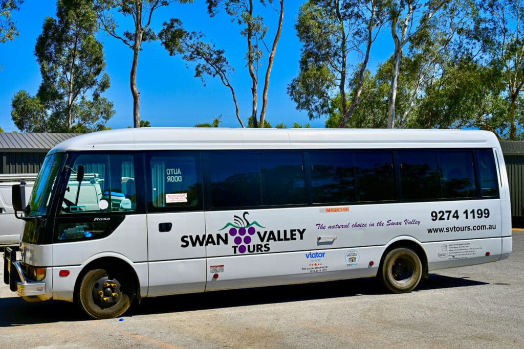 A white coach parked facing the left. It has branding for Swan Valley Wine Tours. Blue sky above and tall green trees
