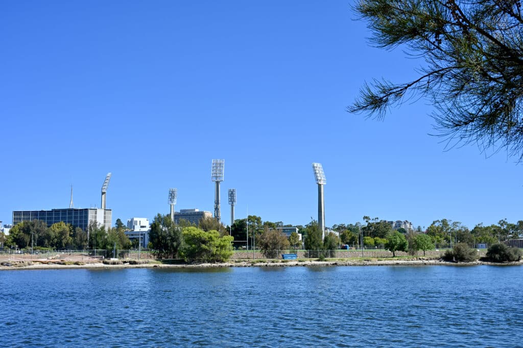 View from a river in the bottom of the frame to a row of trees on the river bank. Above it are the spotlights on tall concrete arms above a sports ground. Blue sky above.