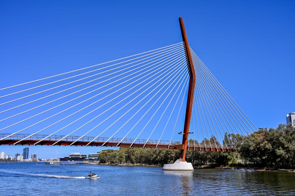 Looking down a river towards a bridge with tall post and white cables radiating from either side of it. trees and buildings behind. Blue sky above.