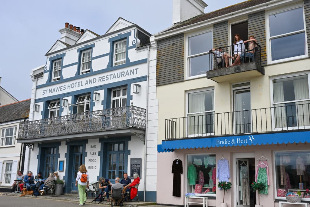 Two buildings fill the frame. A white 3 storey building to the left with the words St Mawes Hotel and Restaurant. It has a wrought iron balcony across the front. Cafe tables on the pavement with people having coffee. A shop building to the right beneath two floors of flats with balconies and people on the top balcony. The shop says Bridie and Bert on the blue awning.