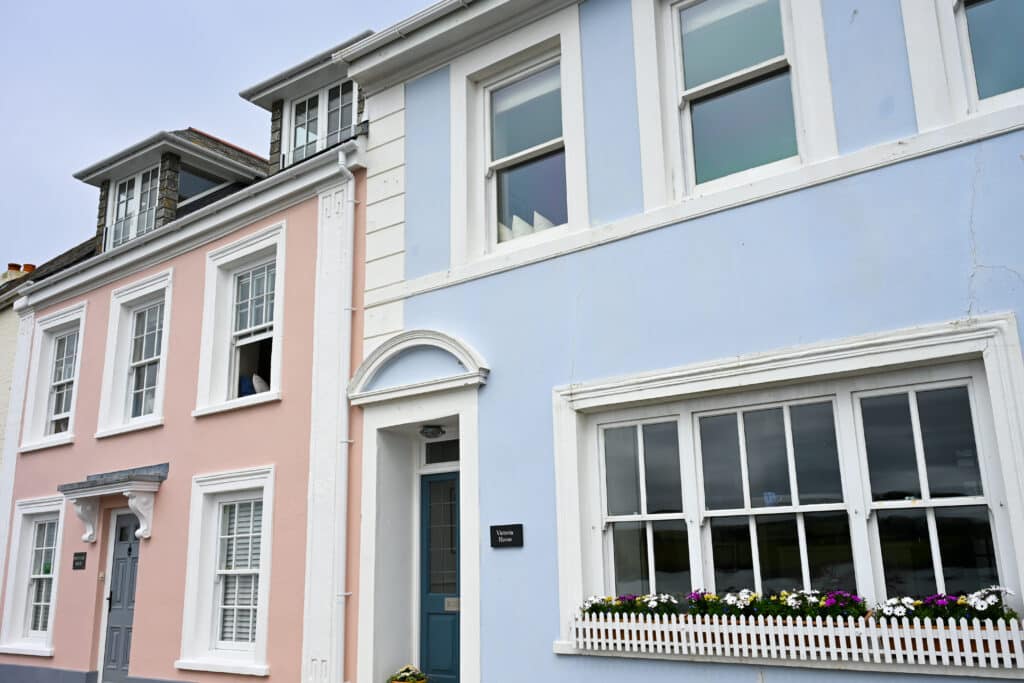 A row of two storey houses runs across the frame. Pink house with white window frames to the let and pale blue painted house to the right. Dormer windows above.