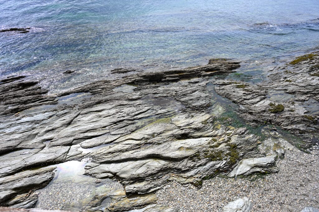 Layers of rock fill the bottom two-thirds of the frame with turquoise water above it. Pebble shore in the bottom right. A frame filled with pattern (rocks) and colour (water)