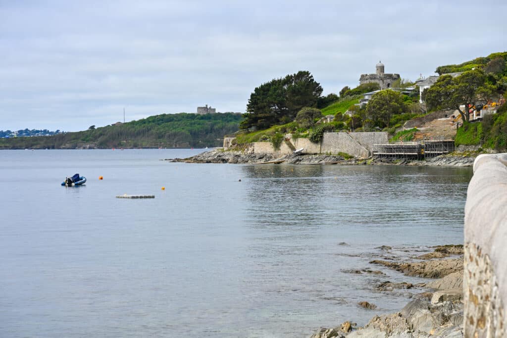 View across a flat body of water to two headlands - one close to the camera with grass, trees on the cliff side and a circular stone castle on top. The second headland is in the distance but also features a circular castle like building and trees. Grey sky above. Stone wall running along the right hand side of the frame.