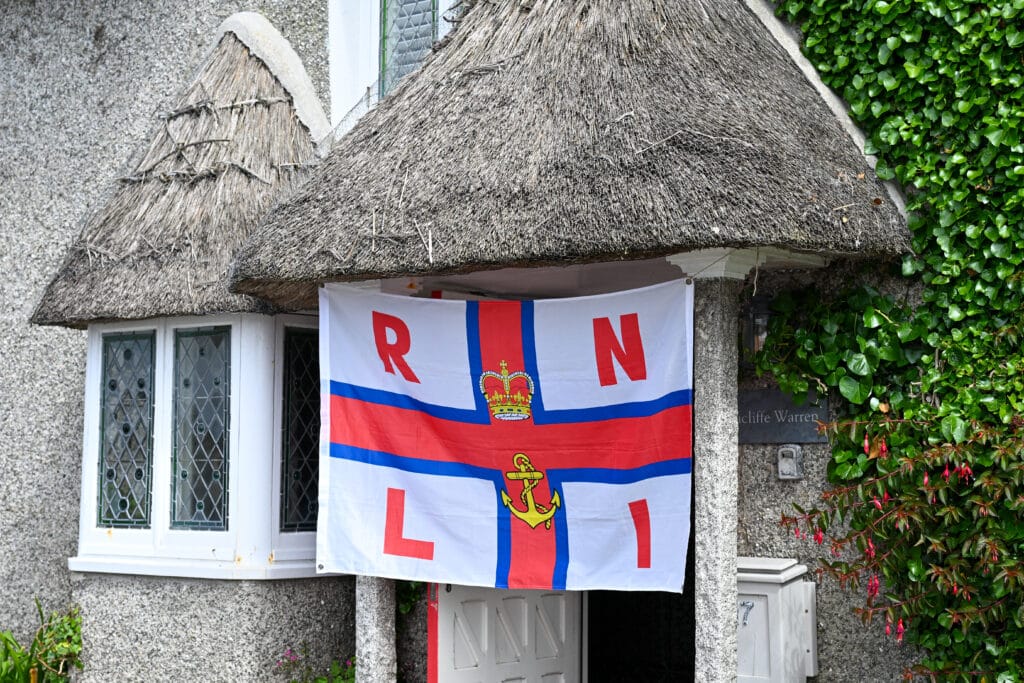 The front of a cottage with thatched roofs above window to the left and door to the right. Some ivy on the right hand side. A large white flag hangs over the door with a Red Cross and the letters RNLI on it.