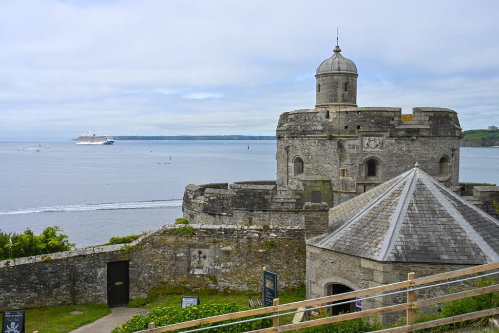 View of a circular stone castle which sits to the right hand side of the frame. It is like a tiered cake. Grey blue water to the left with a cruise ship in the distance. Cloudy sky above.