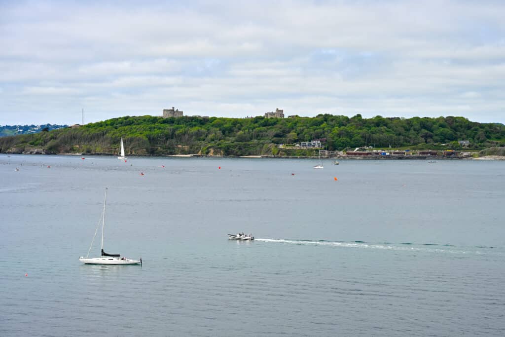 View across a body of water to a strip of green trees on the horizon. There is a castle building on the top of the cliff. Cloudy sky above. Sailing boats and motorboats on the water